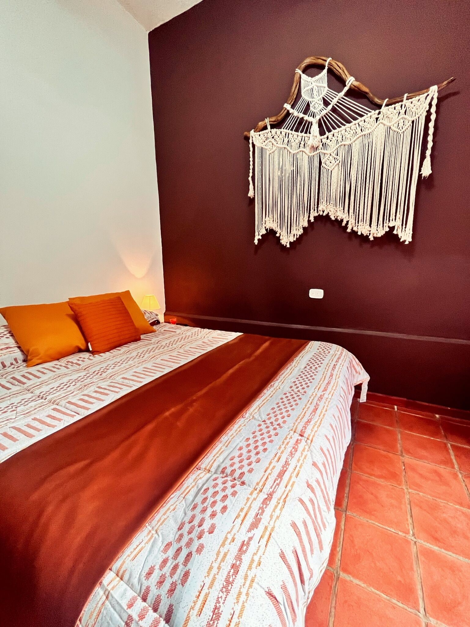 Un lit recouvert d’une couvre-lit à motifs, agrémenté de deux coussins orange, une décoration murale en macramé et un sol carrelé.