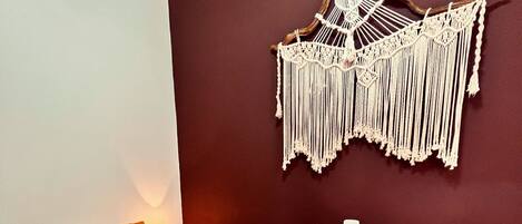 Un lit recouvert d’une couvre-lit à motifs, agrémenté de deux coussins orange, une décoration murale en macramé et un sol carrelé.