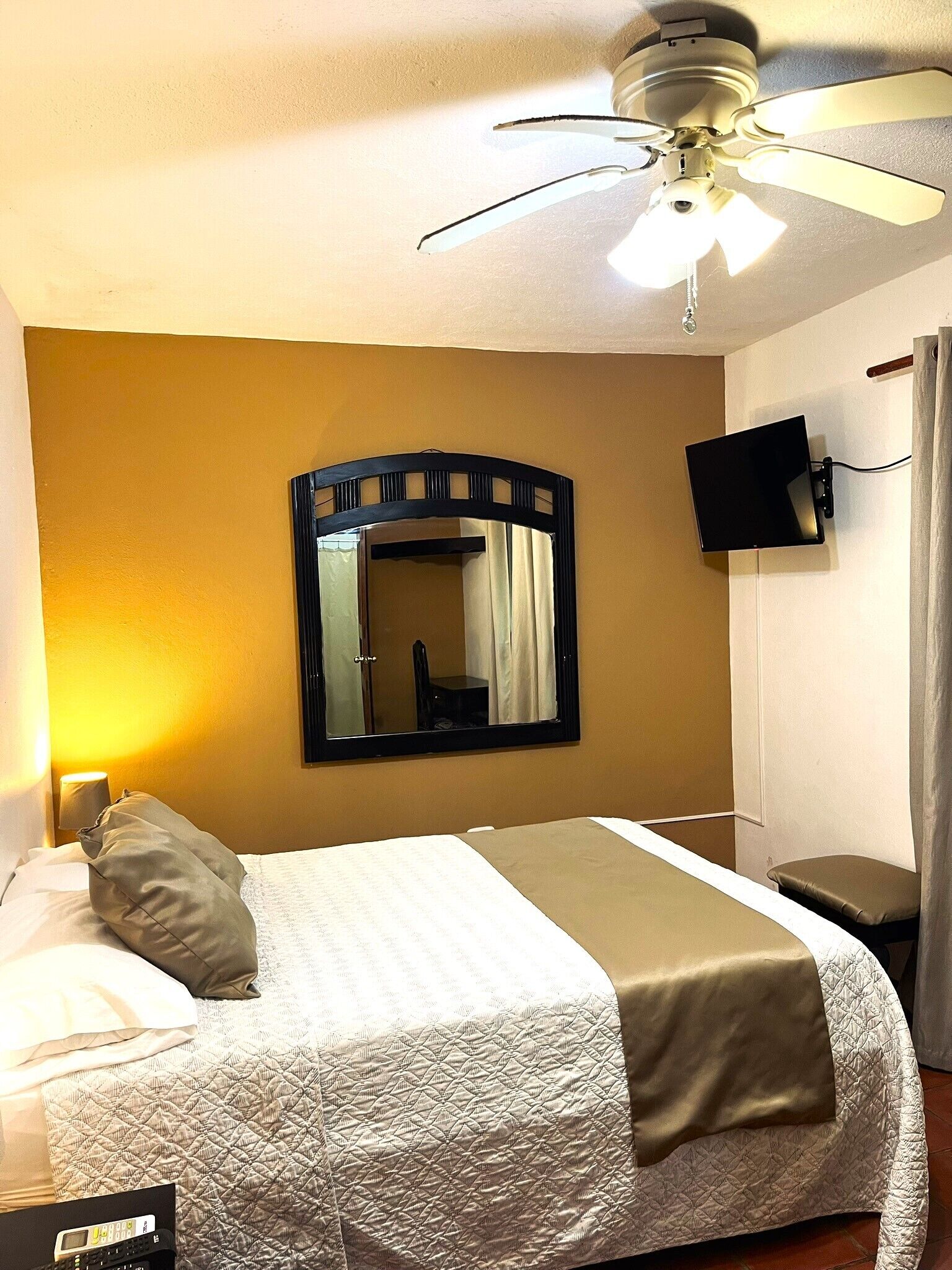 A hotel room with a bed, a ceiling fan, a flat-screen TV mounted on the wall, and a mirror.
