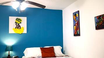 Une chambre à coucher comprenant un lit, un ventilateur de plafond, une table de chevet et deux tableaux accrochés au mur.