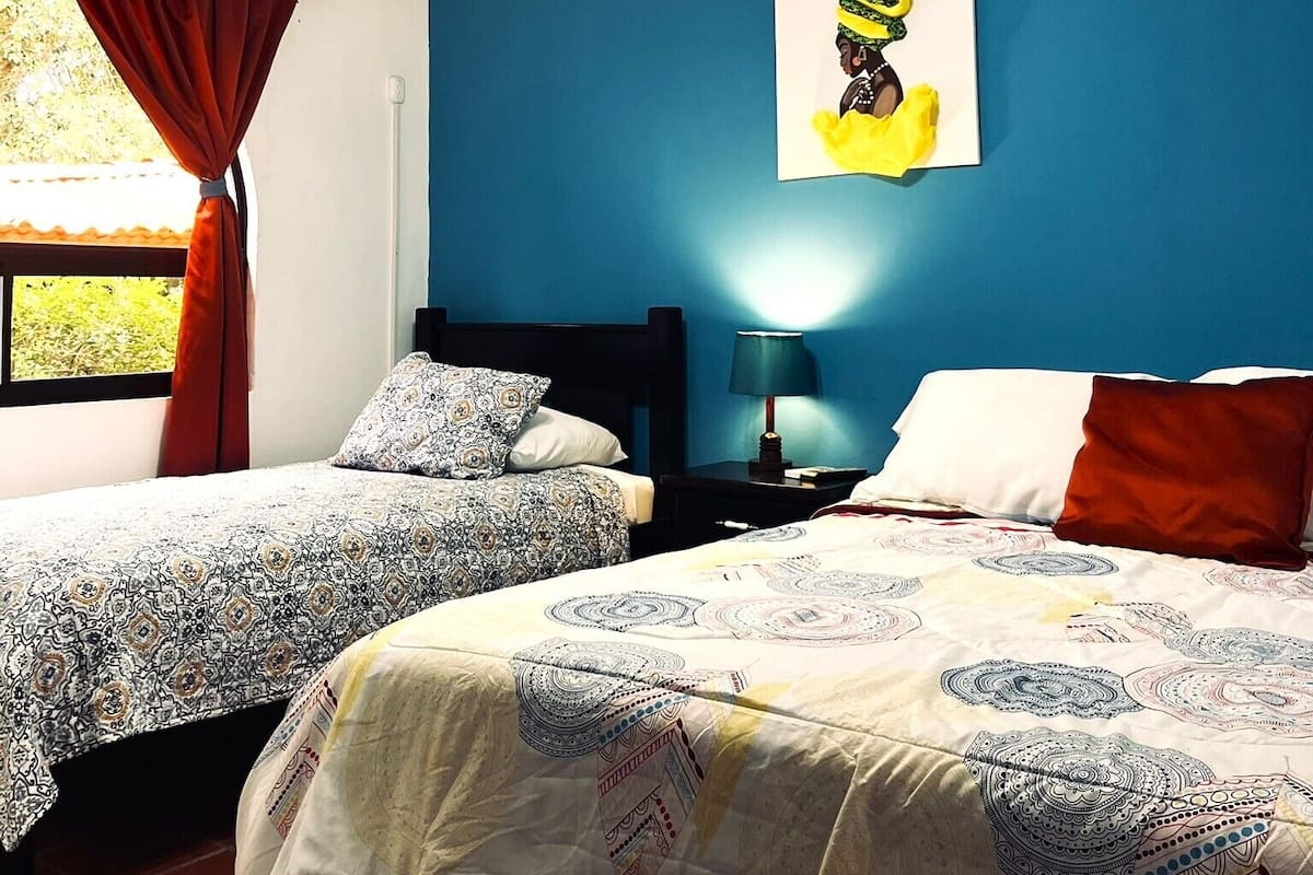 A room with two beds, a ceiling fan, a window with red curtains, and a wall-mounted painting.