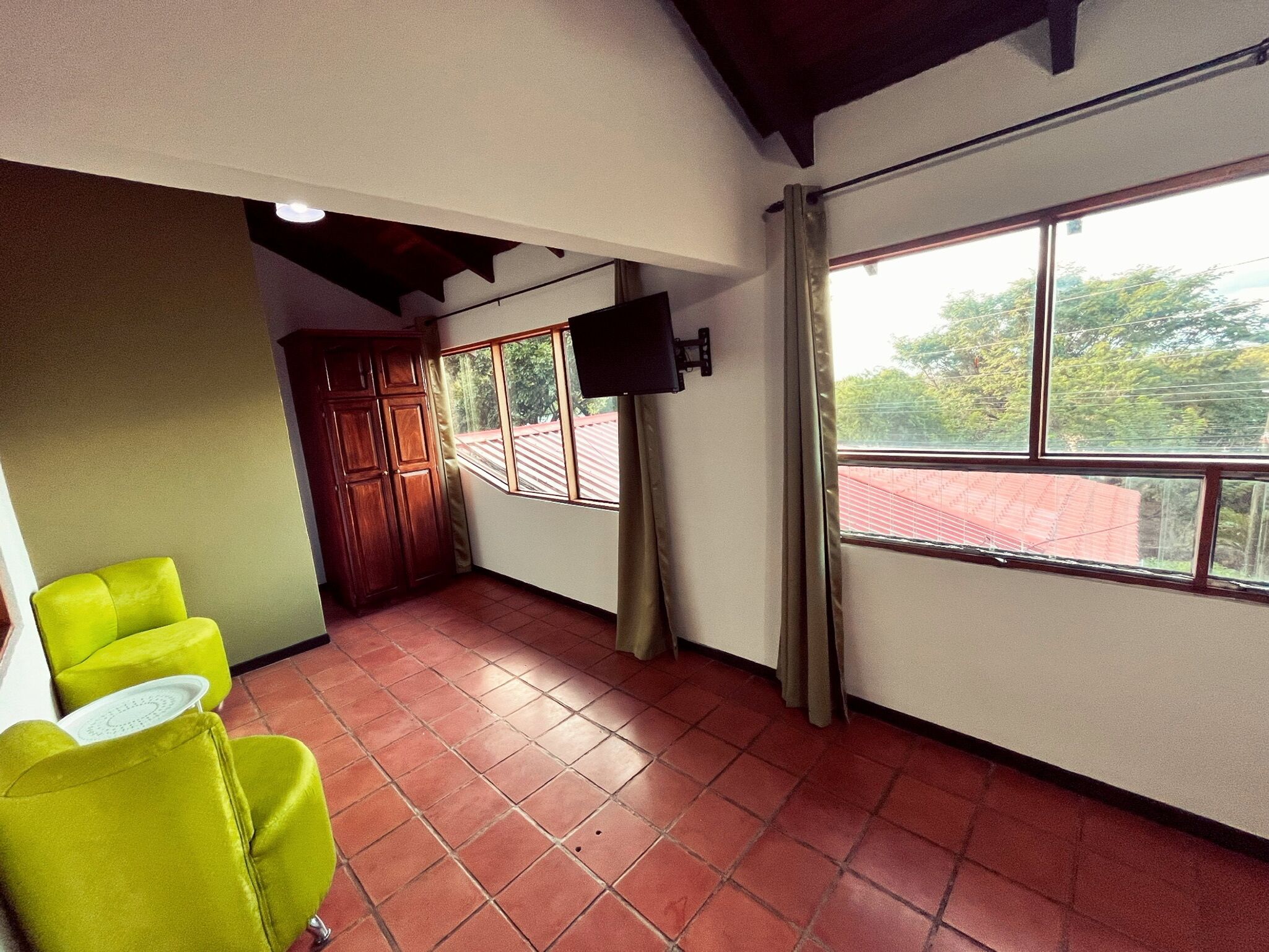 A room with a red-tiled floor, a wooden cabinet, a television mounted on the wall, and a window with curtains.