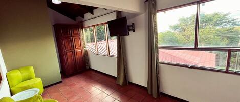 A room with a red-tiled floor, a wooden cabinet, a television mounted on the wall, and a window with curtains.