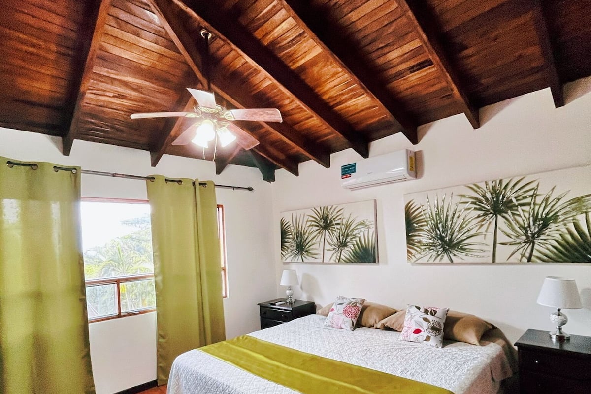 A bedroom with a wooden ceiling, a bed with a green blanket, a nightstand with a lamp, and a window with curtains.