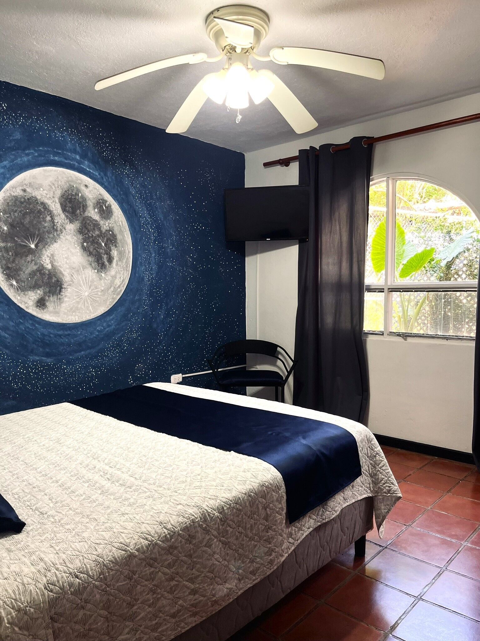 Une chambre à coucher comprenant un lit, un ventilateur de plafond, une fresque murale représentant la lune, une fenêtre avec des rideaux et une chaise.