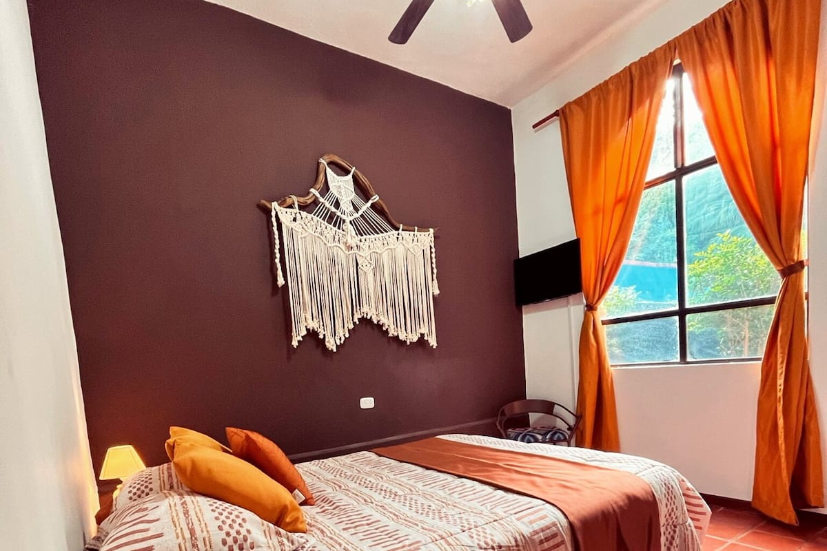 A bedroom with a bed, a ceiling fan, a wall-mounted macrame piece, and a window with curtains.