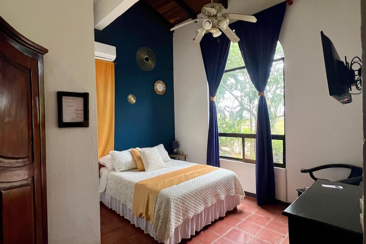 A bedroom with a bed, a desk, a ceiling fan, and a window with curtains.