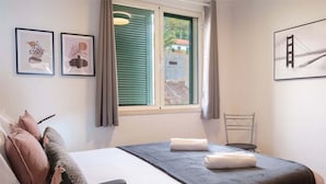 Quarto de hotel com cama, almofadas, janela com persianas, uma cadeira e duas fotografias emolduradas na parede.