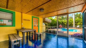A room with a wooden ceiling, a table with benches, a window with blinds, and a view of a swimming pool and greenery outside.