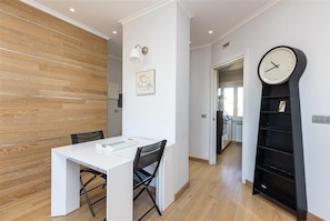 Eine moderne Küche mit Holzböden, einem weißen Esstisch mit Stühlen, einem schwarzen Bücherregal und einer an der Wand befestigten Uhr.