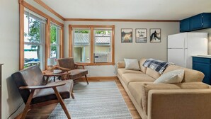 A cozy living room with a sofa, chairs, a small table, and a rug. There are windows with blinds, a refrigerator, and wall art.