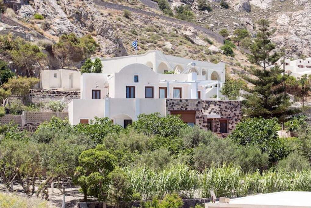 Un edificio bianco con facciata in pietra, circondato da vegetazione e un pendio roccioso.