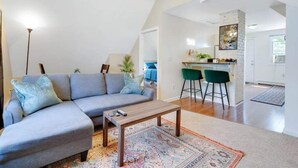 Ein modernes Wohnzimmer mit einem grauen Ecksofa, einem Holztisch und einem gemusterten Teppich. Im Hintergrund ist eine Küchenzeile mit einem Barhocker und einem grünen Stuhl zu sehen.