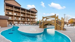 A pool area with a wooden bridge, lounge chairs, and a multi-story building with balconies.