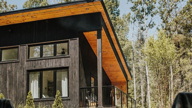 Una cabaña moderna con terraza de madera, ventanas amplias y zona con fogata.