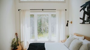 Un dormitorio con cama, una guitarra, una planta y vistas a árboles a través de una ventana.