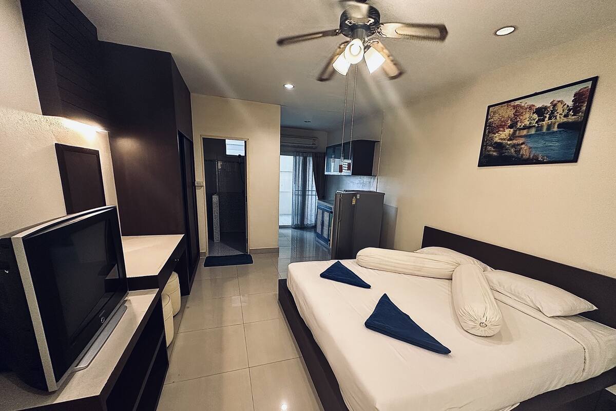 A hotel room with a bed, a TV, a ceiling fan, and a view of the kitchen and bathroom.