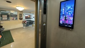 An elevator interior with a digital screen displaying a cityscape, a reception desk, and a green mat on the floor. - Navanakorn Aomsin Hotel near Big C Super (Khlong Luang)