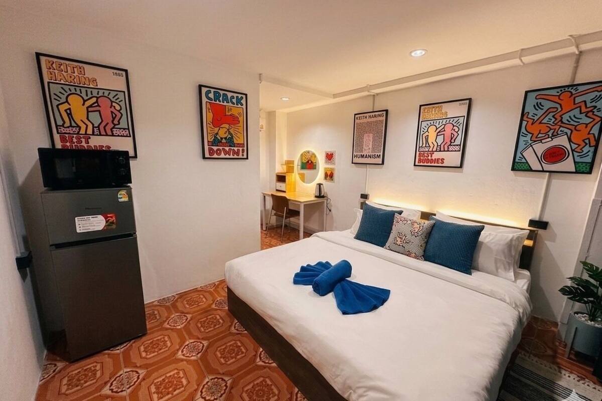 A hotel room with a bed, a desk, and a refrigerator. There are posters on the walls.