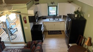 A compact kitchen with white cabinets, a stove, and a sink. There is a dining area with a table and chairs, and a view of an outdoor deck. - The Walnut at Hamilton Cove ~ Cabin Nineteen ~ Pet friendly ~ Lakefront (Royal)