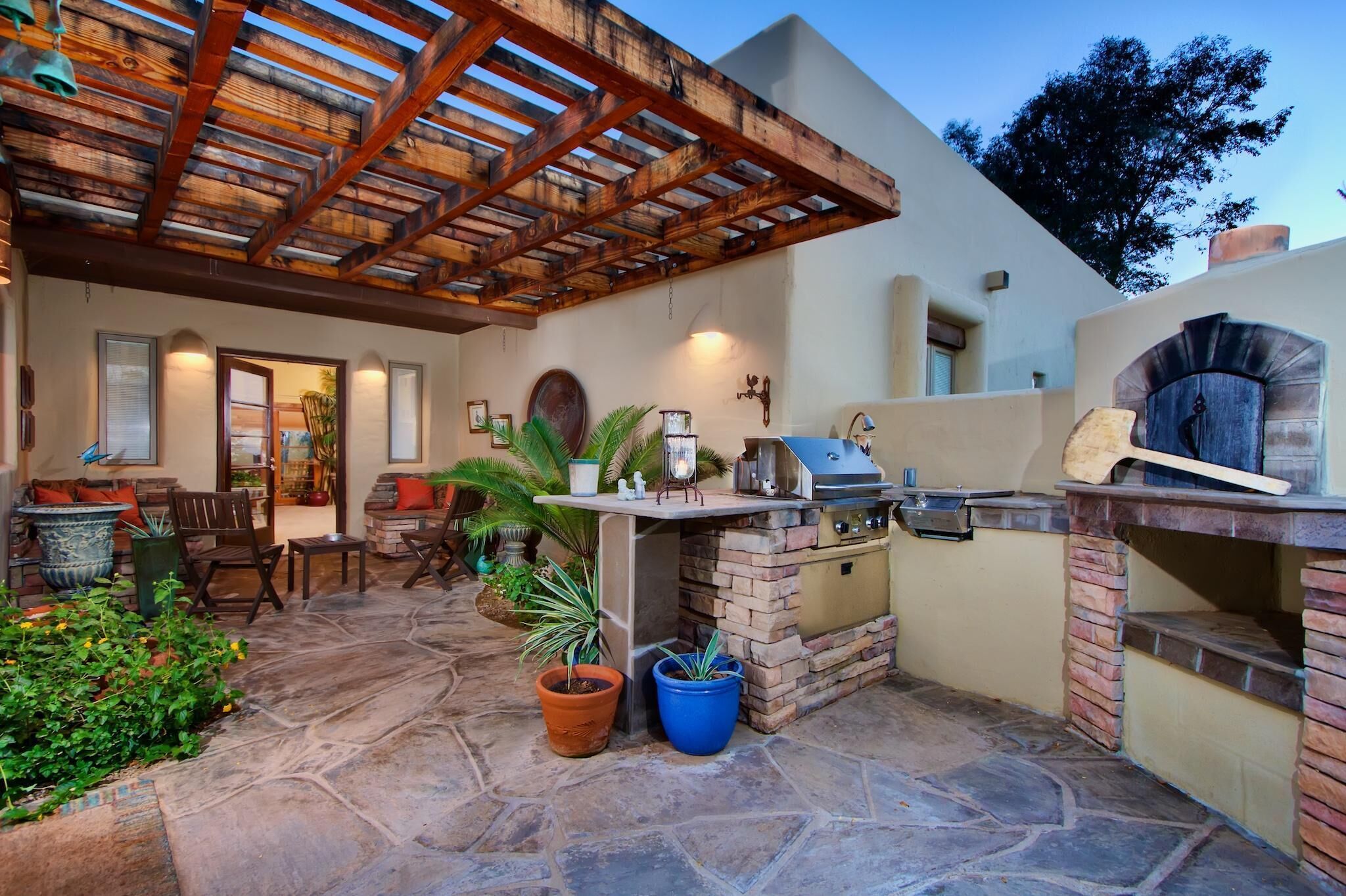 An outdoor patio with a stone floor, a wooden pergola, a grill station, and potted plants.