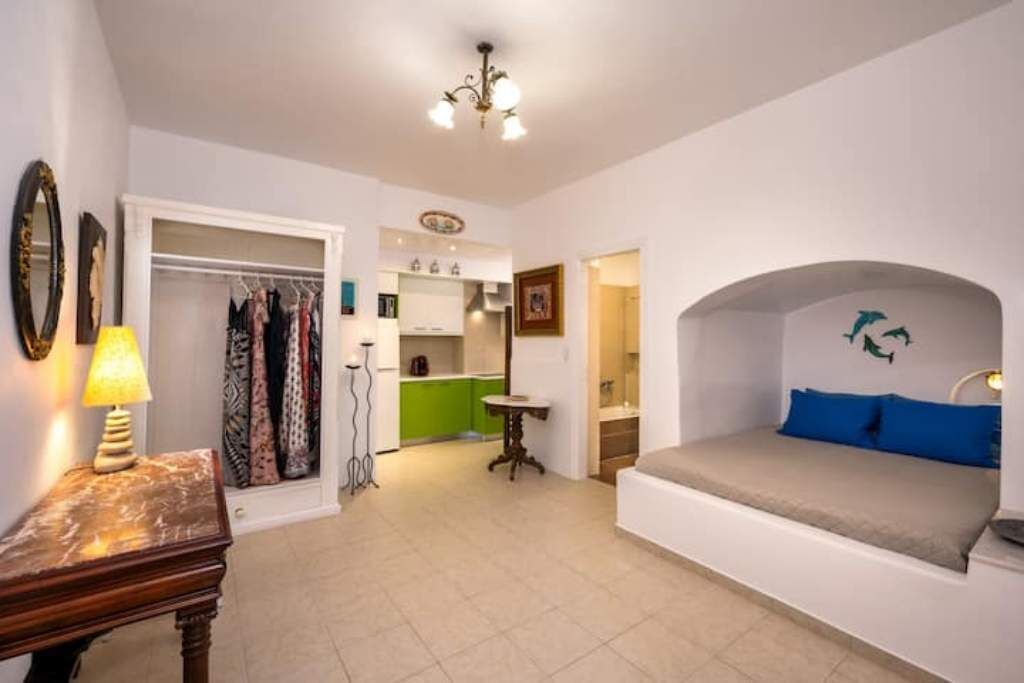 Ein kleines Zimmer mit Bett, einem Tisch, einer Lampe, einem Kleiderschrank und einer Küchenzeile.