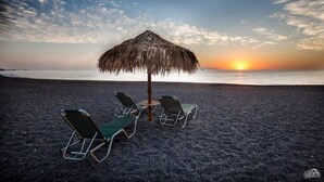 A beach scene with a thatched umbrella and four lounge chairs facing the ocean at sunset. - MOSCHATOS BEACH HOUSE (Καμάρι)