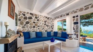 A living room with a stone wall, blue sofa, white coffee table, and a fireplace. - MOSCHATOS BEACH HOUSE (Καμάρι)