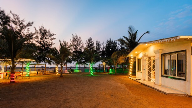 A sandy outdoor area with palm trees, a small building with a porch, and a view of the ocean. - Treebo Salt Water Beach Resort (Dapoli)