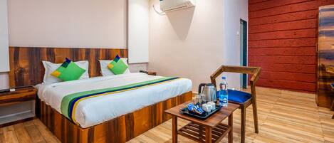 A hotel room with a wooden bed, a bedside table with a tray of toiletries, a red brick wall, and a ceiling-mounted air conditioner.