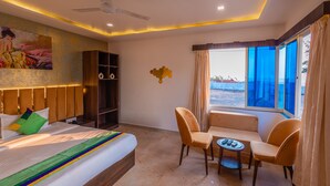A hotel room with a bed, a seating area with two chairs and a table, a ceiling fan, a painting on the wall, and a large window with a view. - Treebo Salt Water Beach Resort (Dapoli)