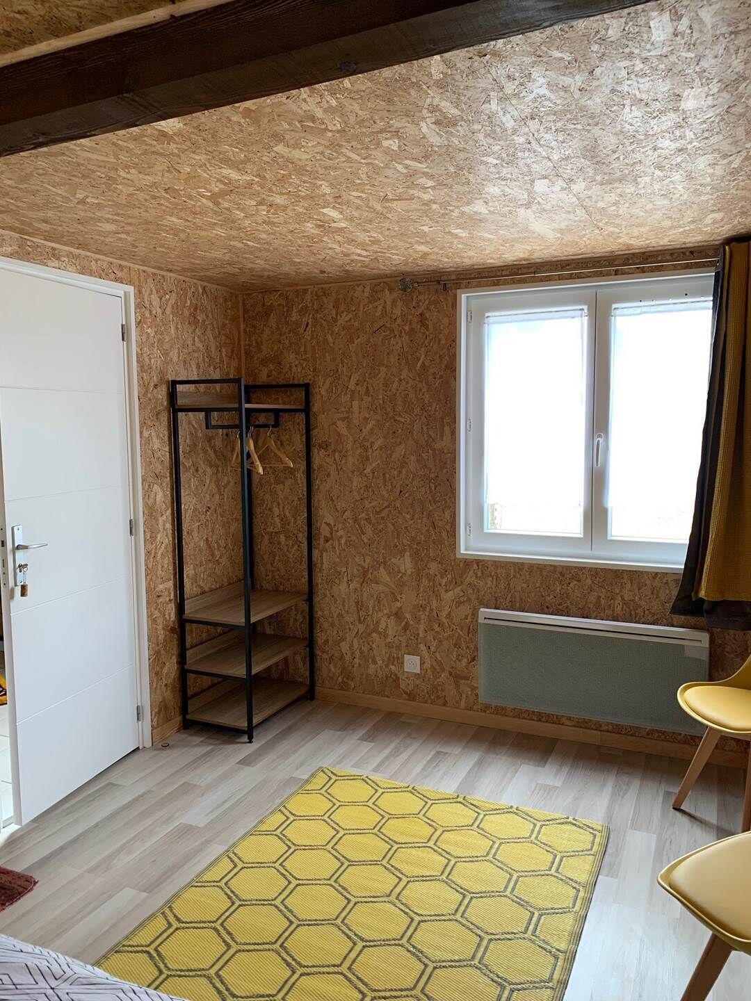 A room with a wooden wall, a window, a radiator, a yellow hexagonal rug, and a yellow chair.