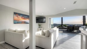 A modern living room with two sofas, a flat-screen TV, and a large window offering a city view. - Ocean View Poolside Retreat
 (Oceanside)