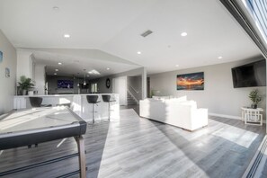 A modern living room with a pool table, a white sofa, and a kitchen area in the background. - Ocean View Poolside Retreat
 (Oceanside)