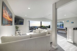 A modern living room with a large sofa, a TV mounted on the wall, and a view of the outdoors through a sliding glass door. - Ocean View Poolside Retreat
 (Oceanside)