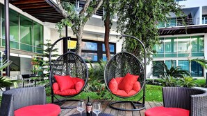 An outdoor seating area with wicker chairs and a table, two hanging chairs with red cushions, and a bottle of wine.