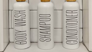 Three white bottles with wooden caps labeled 'BODY WASH,' 'SHAMPOO,' and 'CONDITIONER' against a tiled wall. - The Gate  -   Studio (Skaneateles)