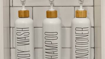 Drei weiße Flaschen mit Etiketten für Duschgel, Shampoo und Conditioner, jeweils mit Holzdeckel, vor einer gefliesten Wand.
