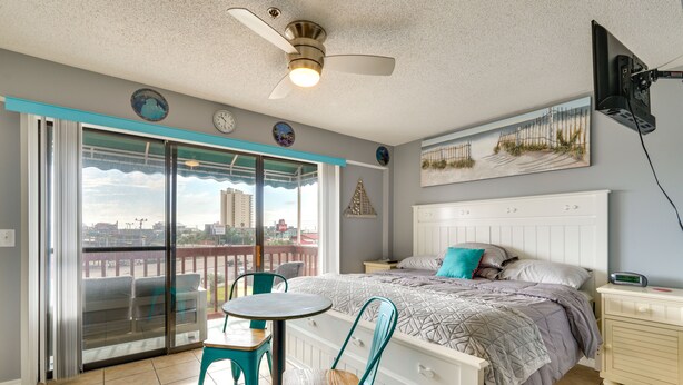 A bedroom with a bed, a nightstand, a table with chairs, a ceiling fan, a window with a view of buildings, and a painting on the wall. - Panama City Beach Getaway - Walk to Beaches! (Panama City)