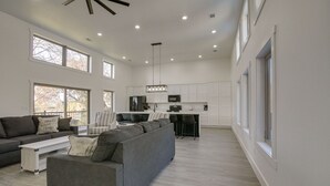 Un amplio espacio de sala con un sofá seccional gris grande, una mesa de centro blanca y una silla a rayas. Se ve una cocina con gabinetes blancos y una zona de bar al fondo.