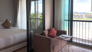Une chambre d’hôtel avec un lit, un canapé, une petite table avec un livre, et de grandes fenêtres donnant sur un immeuble et des espaces verts.