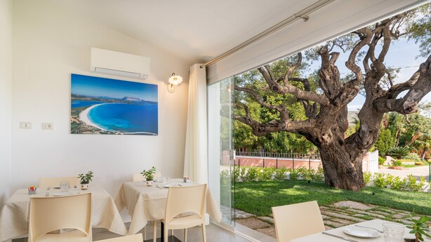 A dining area with tables set for two, a large tree visible through the glass door, and a painting of a coastal scene on the wall. - Residenza L'Alzoni (San Teodoro)