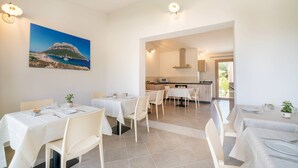 A dining area with tables set for two, a kitchen in the background, and a painting of a coastal landscape on the wall. - Residenza L'Alzoni (San Teodoro)