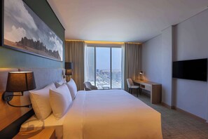 A hotel room with a large bed, a desk, a chair, a TV, and a view of the city through large windows.