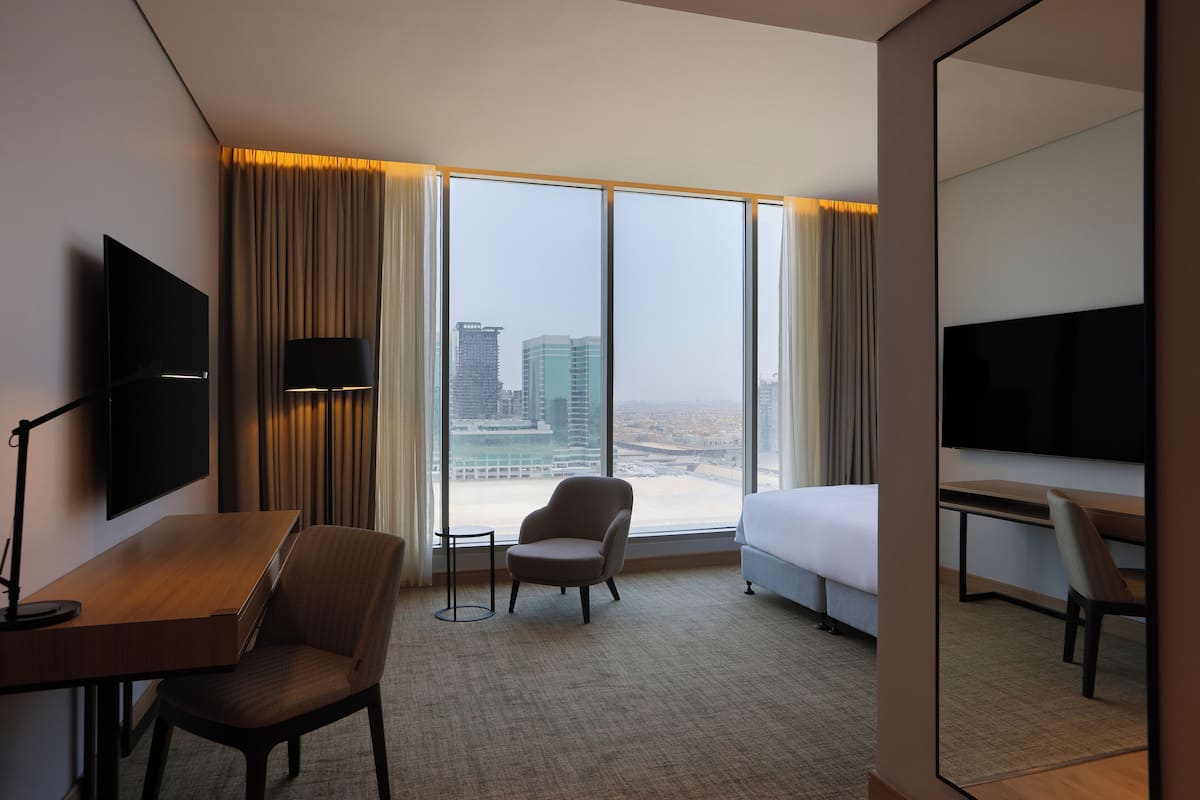 A hotel room with a large window, a bed, a desk, a chair, and a TV.
