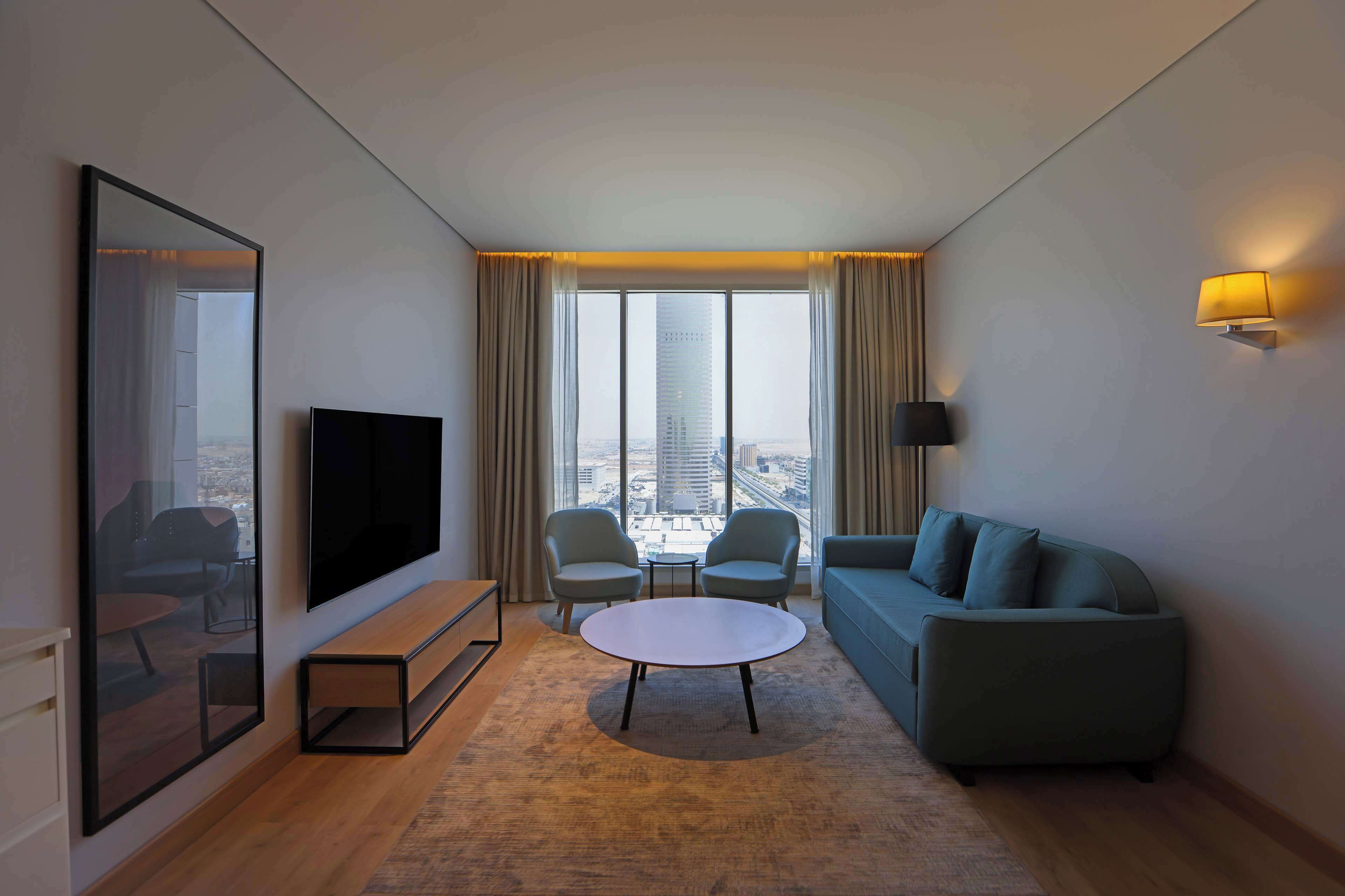 A modern hotel room with a large TV, two armchairs, a sofa, a round coffee table, and a view of a cityscape through large windows.