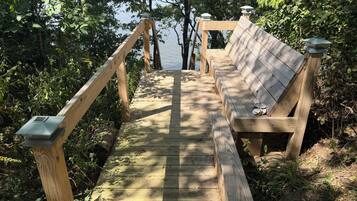Una terraza de madera que lleva a un banco con vistas a un cuerpo de agua, rodeada de follaje denso.