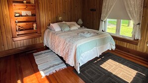 Un dormitorio con paneles de madera, una cama, una mesita de noche, una lámpara y un ventanal con cortinas.