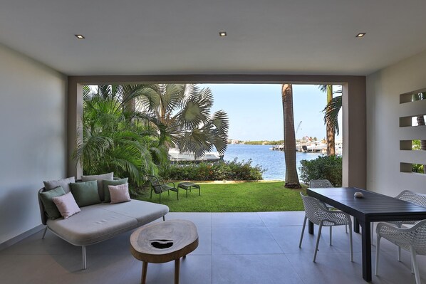 A modern living room with a sofa, dining table, and chairs, overlooking a pool and greenery.