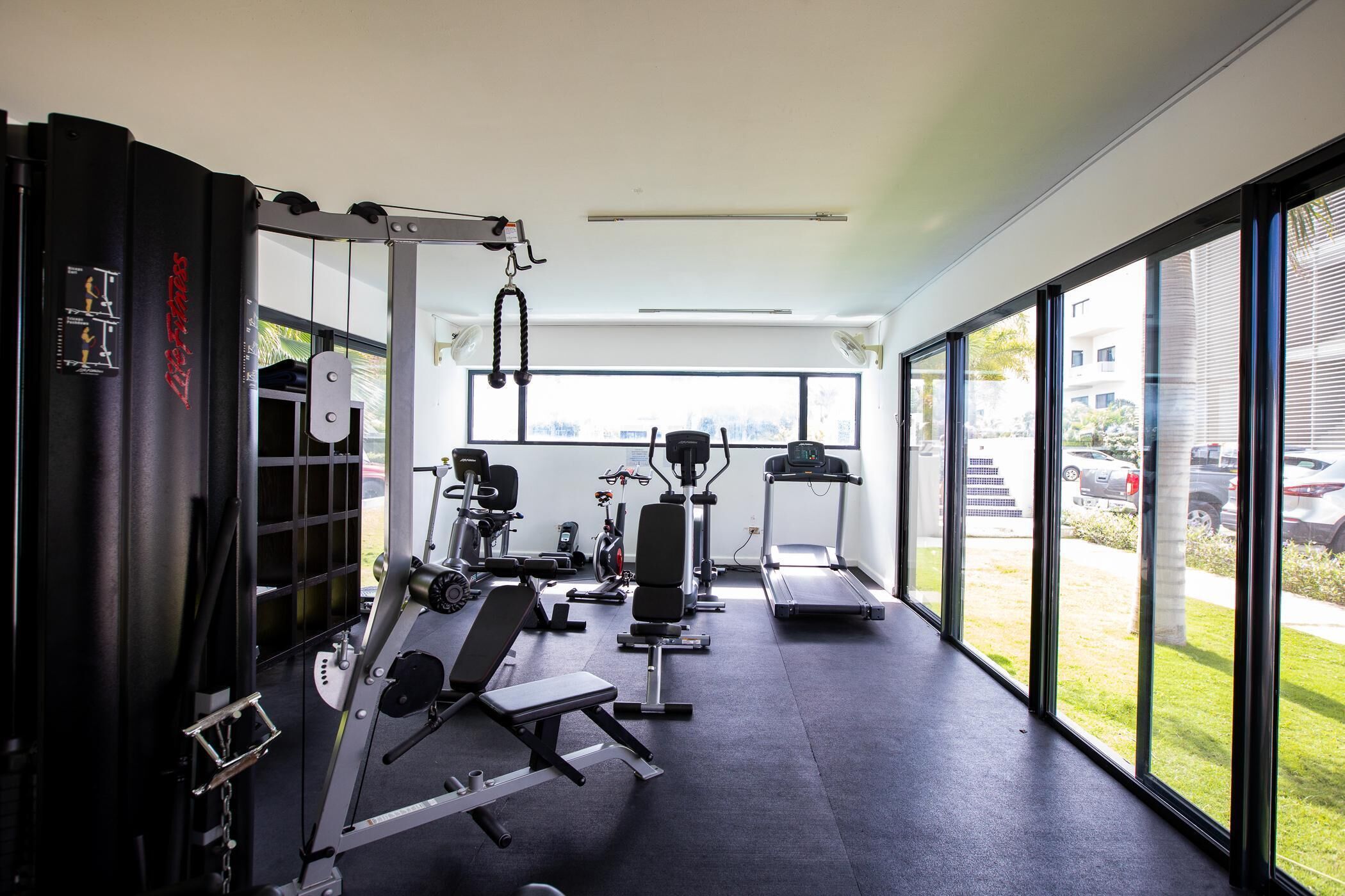 A modern gym with various exercise equipment, large windows, and a view of the outside.